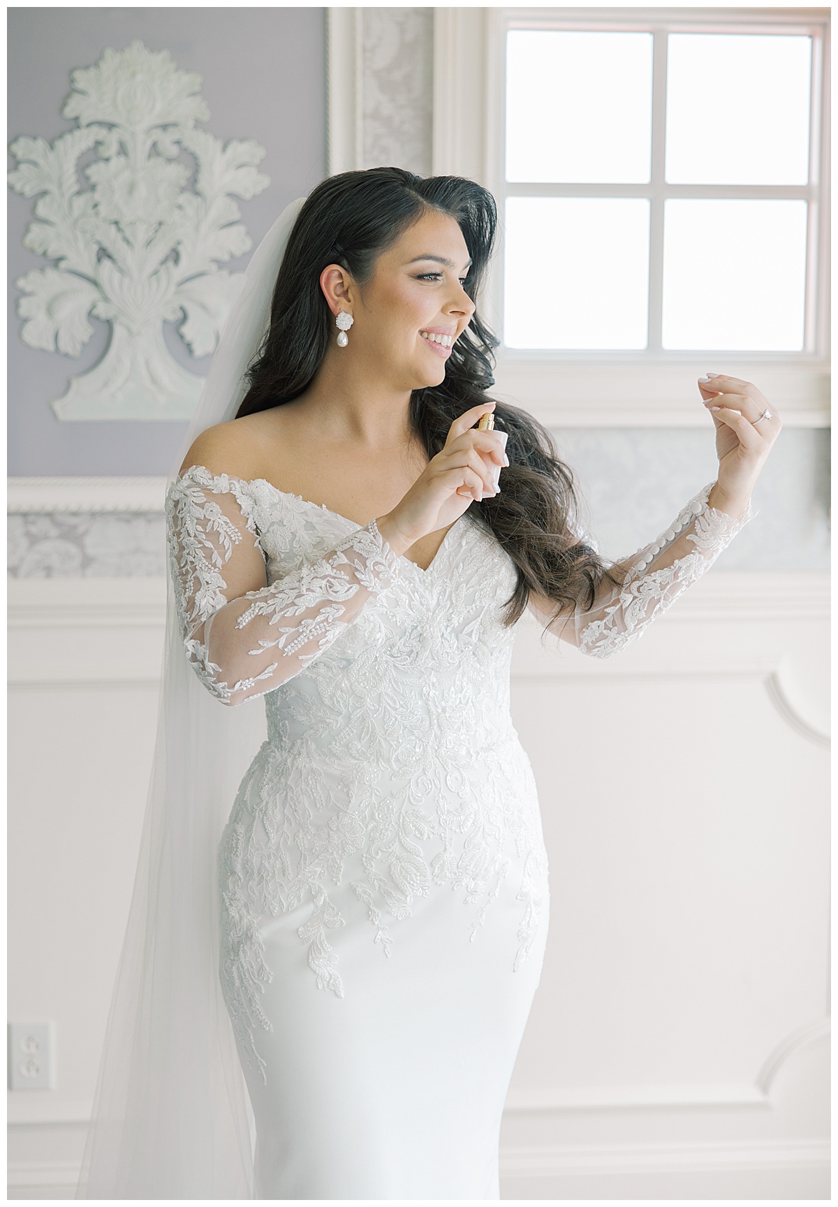Bride spraying perfume on wedding day with lace sleeved dress. 