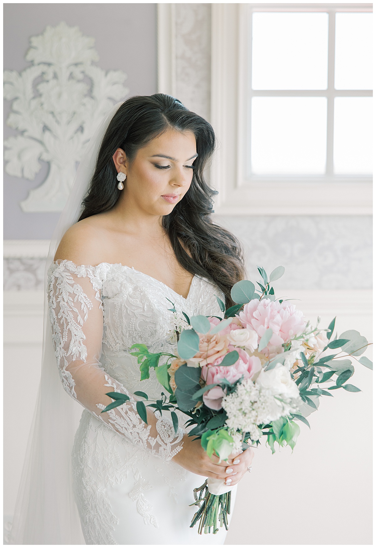 Bride with lace sleeves looking at bouquet with greenery and pink peonies. 
