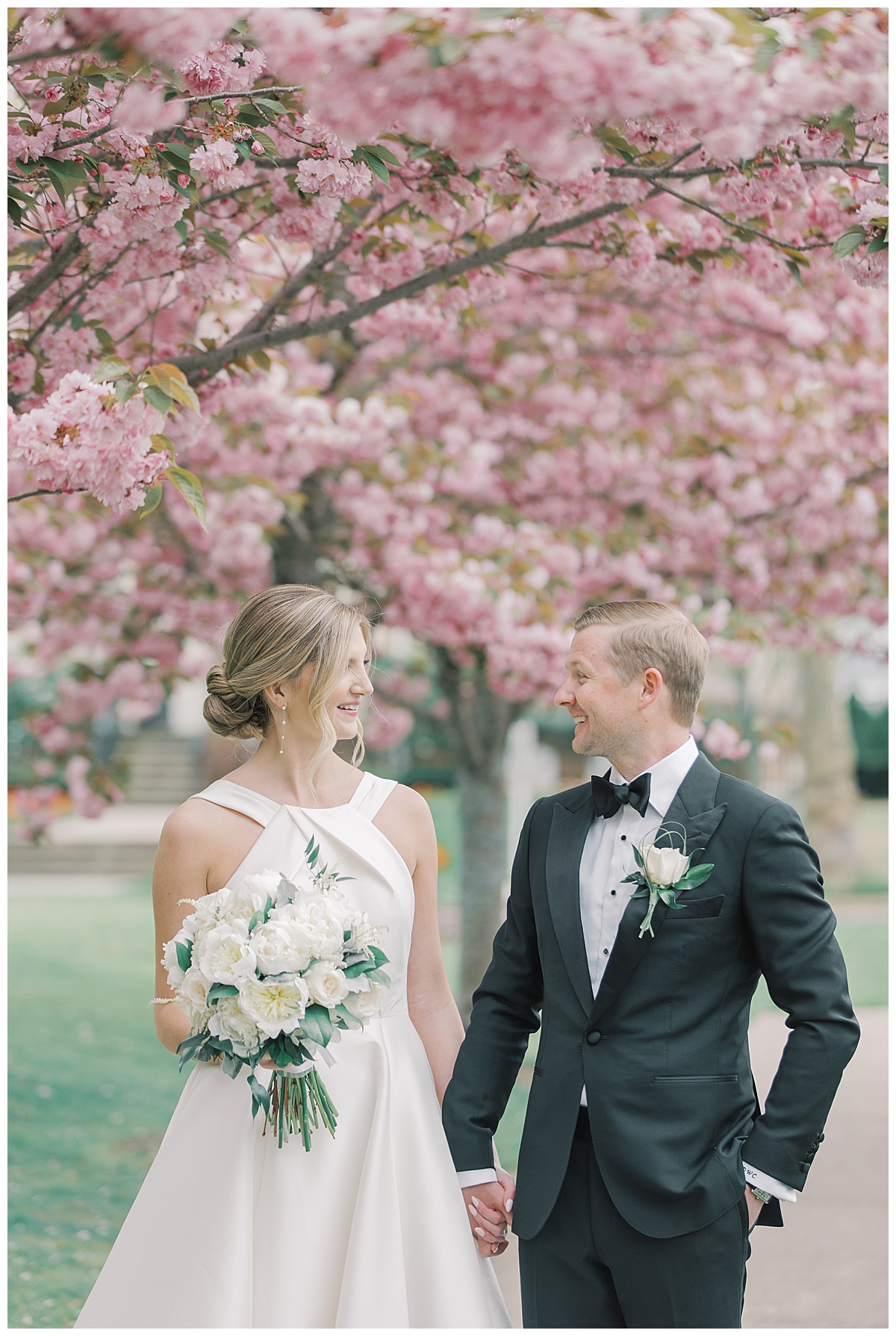 Bride and groom walk while holding hands with cherry blossoms behind them at The Mill Lakeside Manor. 