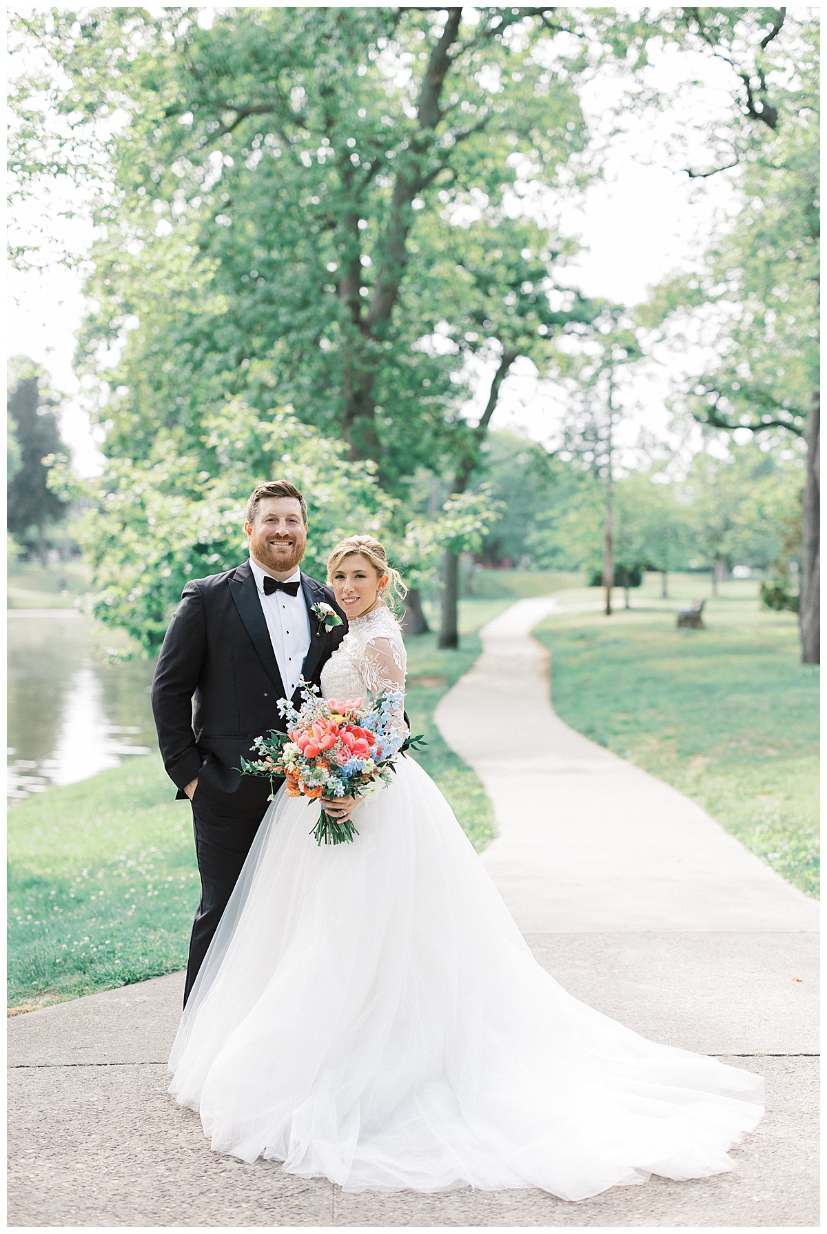 Bride and groom smile together with colorful wildflower bouquet in park with lake.